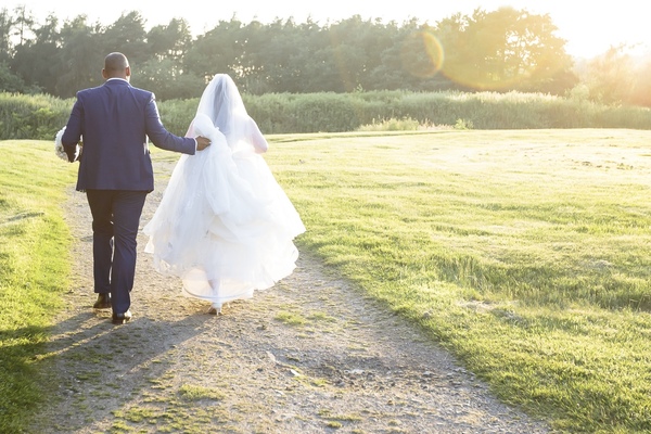 Katie and Manni walk into the sunset. Manni holds both the bouquet and the train of Katie's dress.